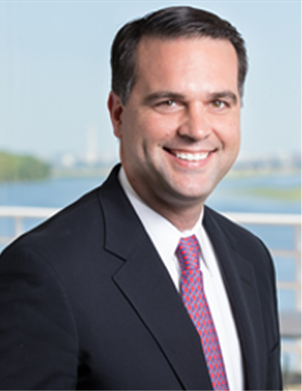 A smiling man in a suit and tie stands outdoors with a river and city skyline in the background.