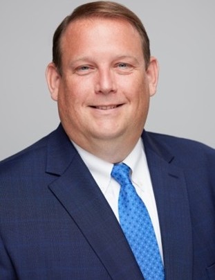 A professional headshot of a man in a navy suit, white shirt, and blue tie, smiling against a light gray background.